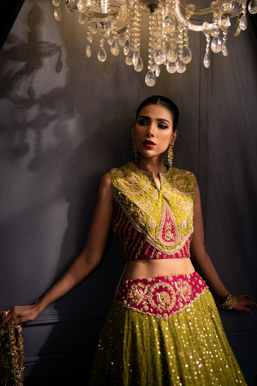 Zohra designer lehenga featuring an embellished choli, paisley motifs, and a flowing olive green skirt and with a hot pink embroidery patch on the hem of the lehenga .
